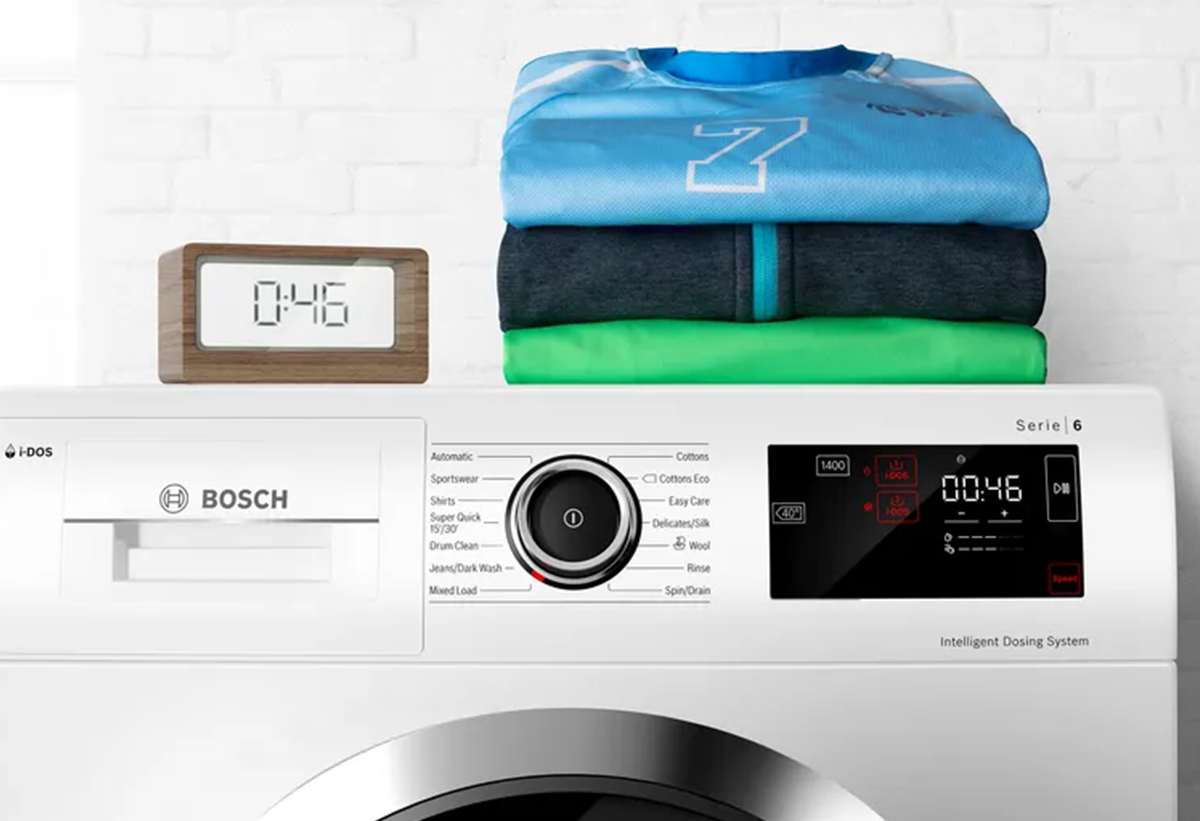 Απεικονίζεται το πλυντήριο ρούχων Bosch  WGG042L9GR πάνω στο οποίο είναι τοποθετημένες διπλωμένες μπλούζες σε διάφορες αποχρώσεις.